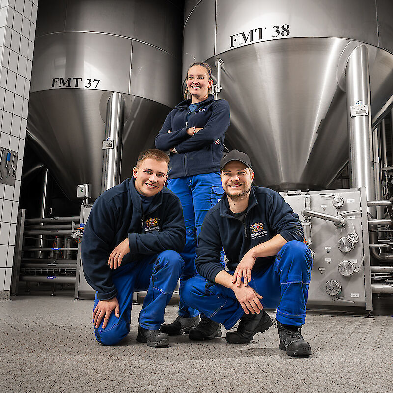 Drei Mitarbeiter in blauer Arbeitskleidung posieren lächelnd vor großen Stahltanks in einer industriellen Brauerei-Umgebung.