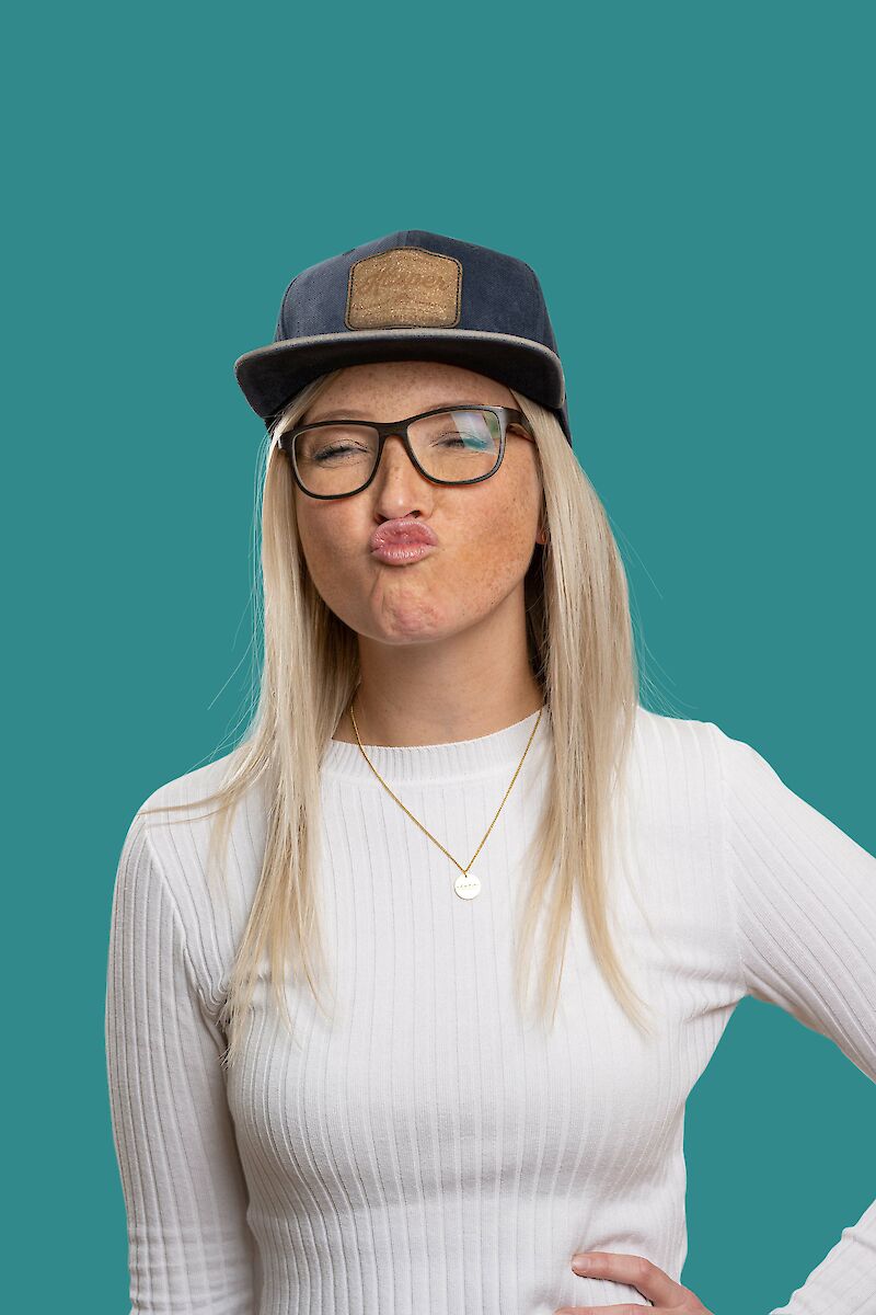 Blonde Frau mit Cap, Brille und Kussmund-Geste vor türkisfarbenem Hintergrund.