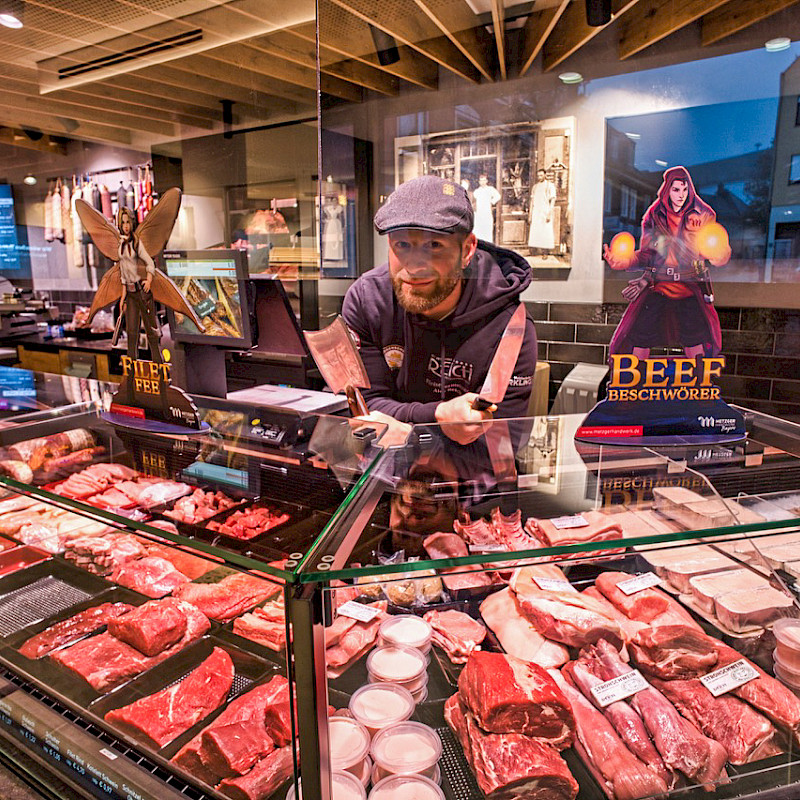 Metzger hinter einer Theke voller Fleischwaren, lächelnd und mit einem Messer in der Hand. Auf der Theke stehen Werbefiguren, die für 'Beef Beschwörer' und 'Filet Fee' aus dem Spiel 'Butcher's Tale' werben