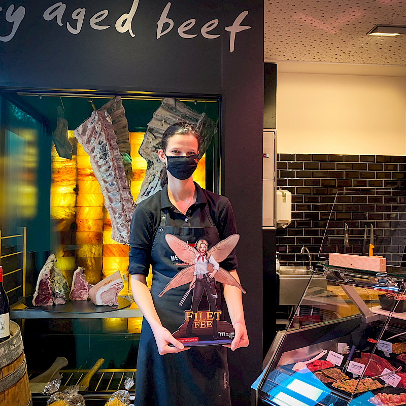Eine Metzgerin mit Gesichtsmaske steht vor einem Dry-Aged-Beef-Schrank und hält eine Werbefigur für 'Filet Fee' aus dem Spiel 'Butcher's Tale'. Im Hintergrund sind verschiedene Fleischstücke zu sehen. Neben der Metzgerin steht ein Fass mit mehreren Flaschen Wein.