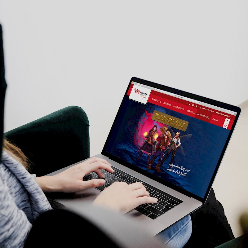 Person sitzt auf einem Stuhl und arbeitet an einem Laptop. Der Bildschirm zeigt die Website von 'Butcher's Tale: Die Wächter des Handwerks' mit drei Fantasy-Charakteren und dem Slogan 'Folge dem Ruf und bewirb dich jetzt!