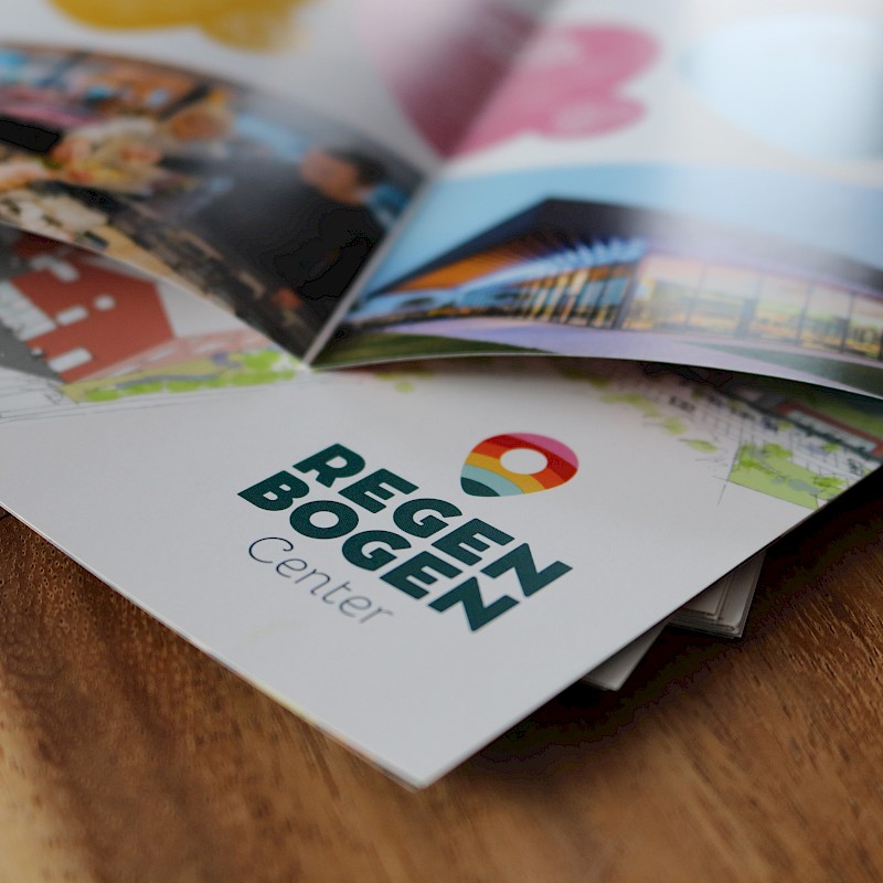 Flyer des Regenbogen Centers auf einem Holztisch, mit Bildern von Aktivitäten und Gebäuden im Hintergrund