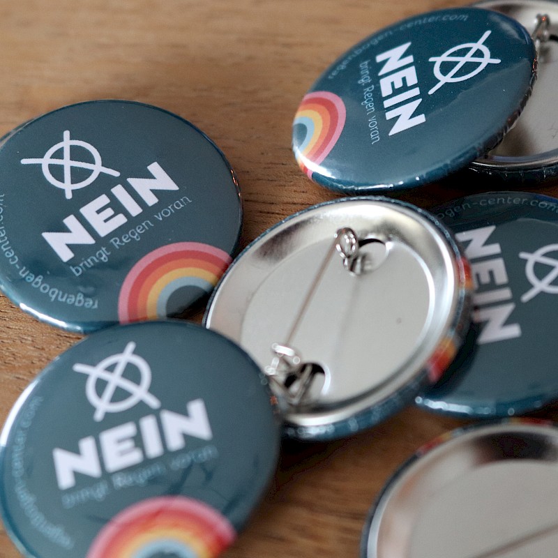 Mehrere Ansteckbuttons auf einem Holztisch mit der Aufschrift 'NEIN bringt Regen voran' und einem Regenbogen-Logo