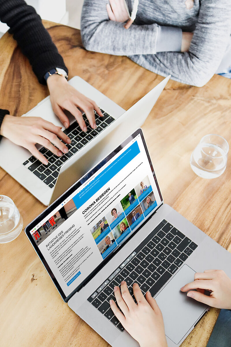 Zwei Personen arbeiten an Laptops, einer zeigt eine Website zum Thema "Corona besiegen