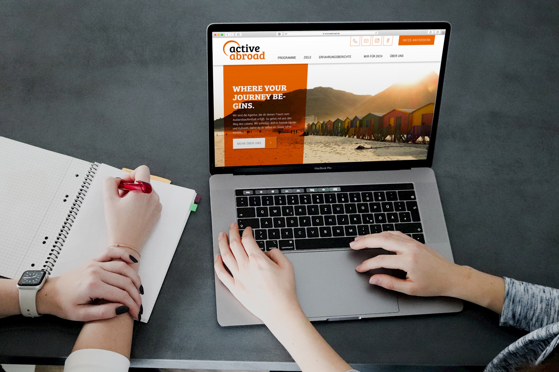 Zwei Personen arbeiten an einem Laptop, der die Webseite von 'active abroad' zeigt. Eine Person tippt auf der Tastatur, die andere schreibt Notizen in ein Notizbuch