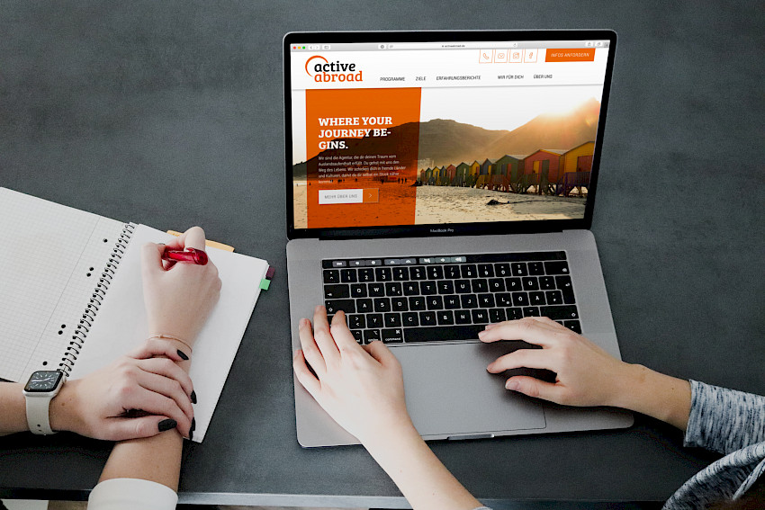 Zwei Personen arbeiten an einem Laptop, der die Webseite von 'active abroad' zeigt. Eine Person tippt auf der Tastatur, die andere schreibt Notizen in ein Notizbuch