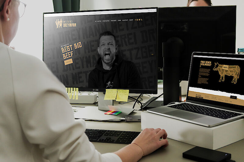 erson arbeitet am Computer, auf dem Bildschirm ist die Website der Metzgerei Dreymann mit dem Slogan 'Best Bio Beef' zu sehen."