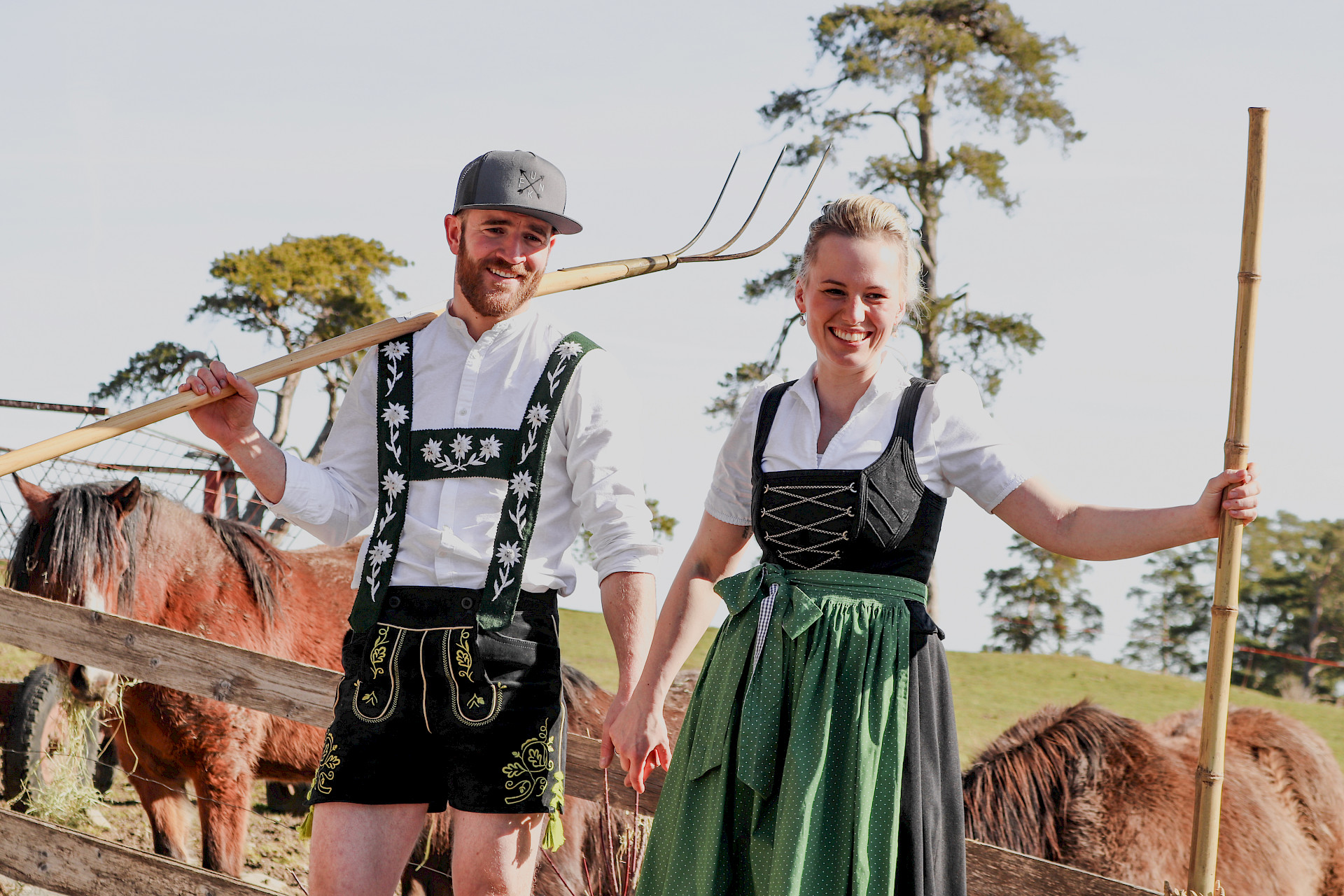 Mann und Frau stehen in Tracht mit Heugabeln vor mehreren Pferden