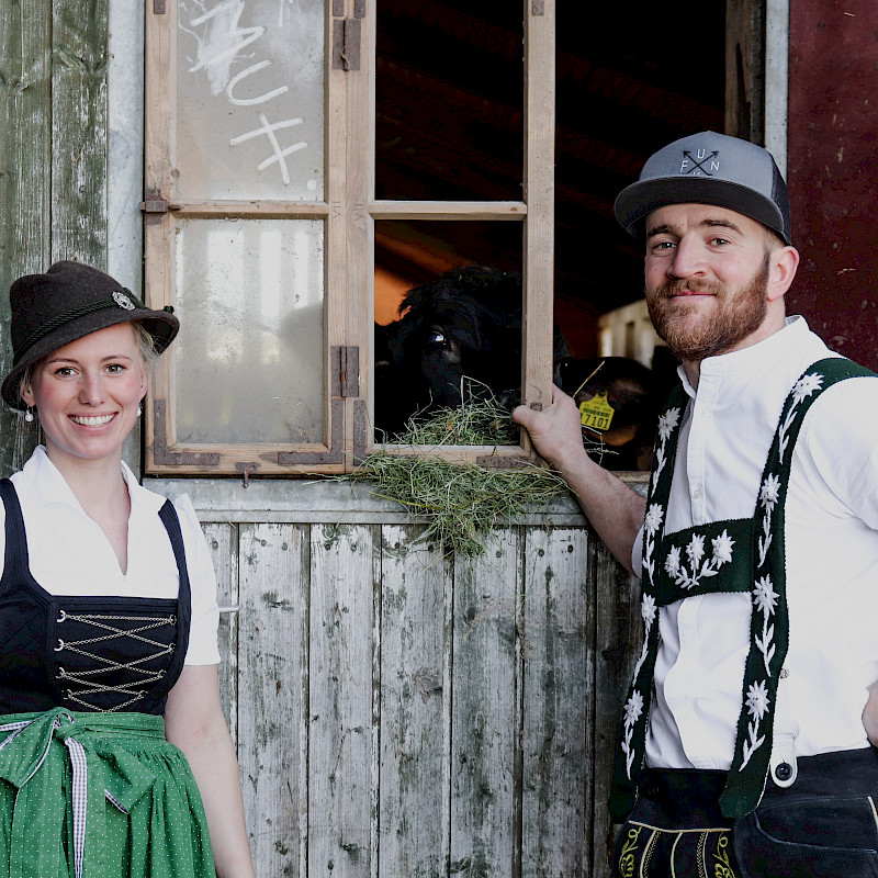 Eine Frau in Dirndl und ein Mann in Lederhosen stehen am Fenster und lächeln.