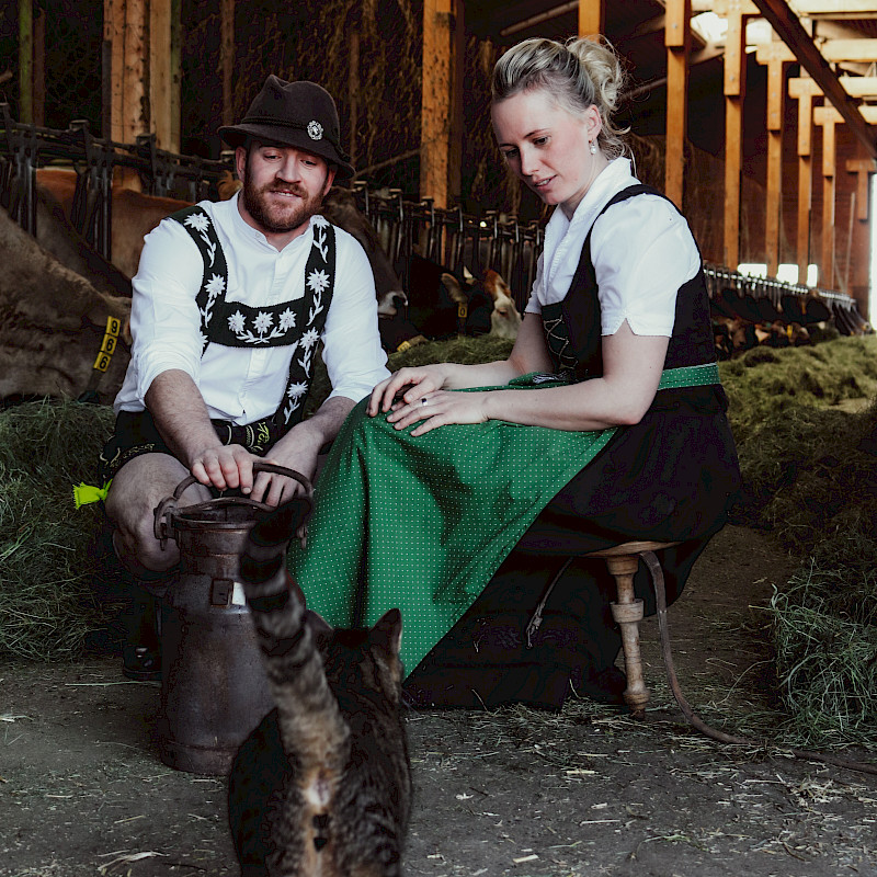 Zwei Personen in traditionellen Dirndl und Lederhosen sitzen in einem Kuhstall und blicken auf eine Katze.