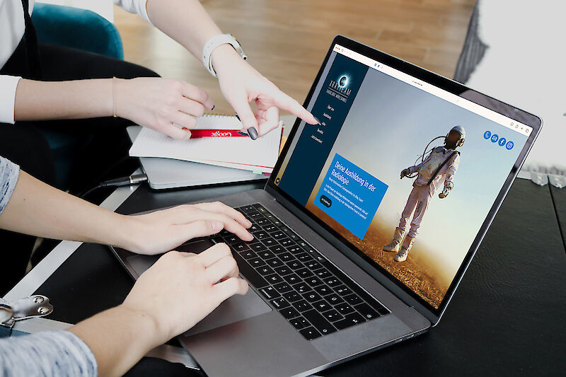 Zwei Personen deuten auf eine Homepage, die auf einen Laptop zu sehen ist.