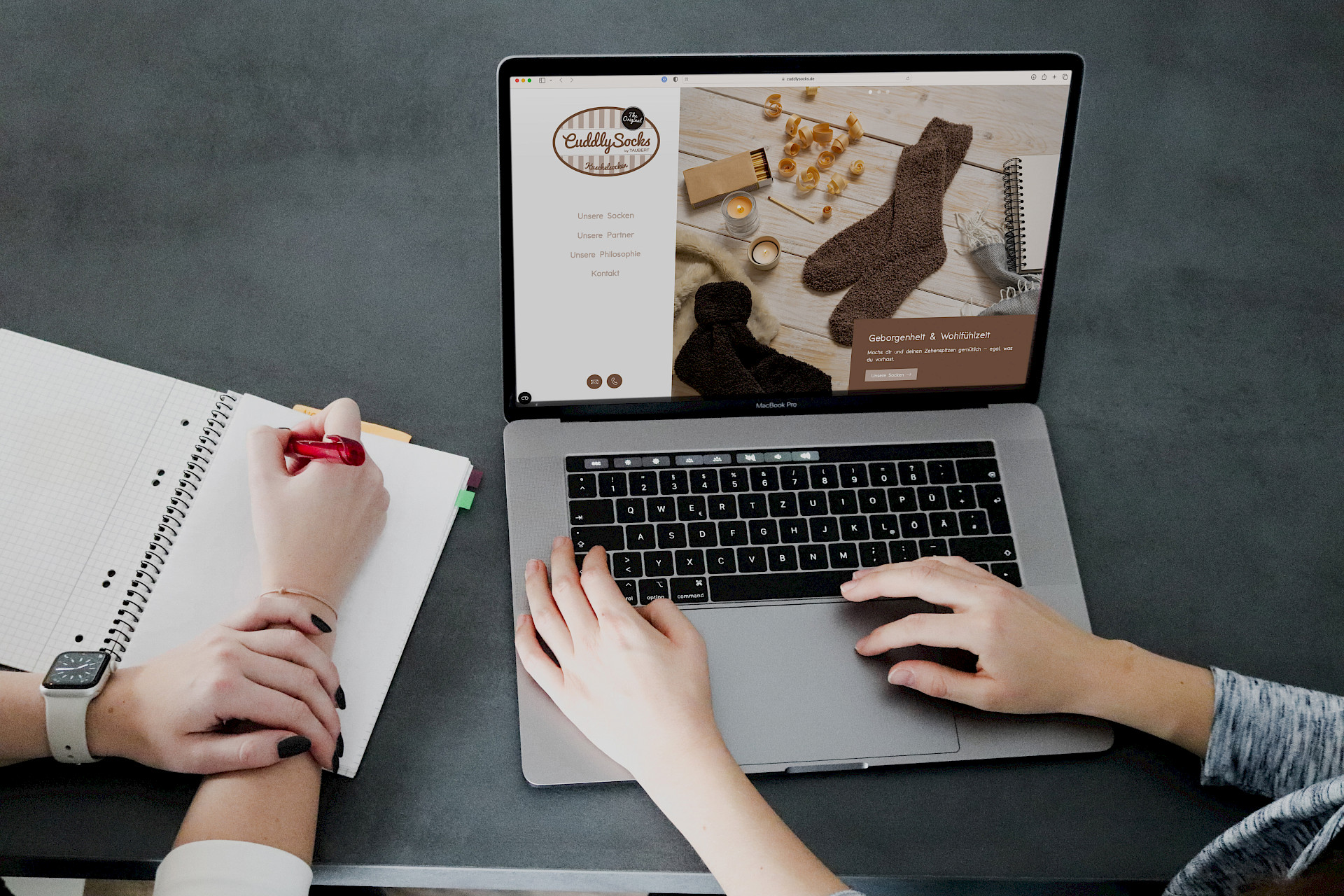 Zwei Personen arbeiten, eine schreibt in ein Notizbuch, die andere nutzt einen Laptop mit der "Cuddly Socks"-Website