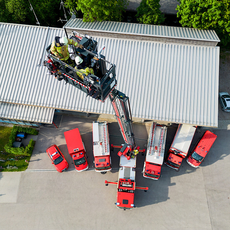 Mehrere Personen stehen in einer Hubarbeitsbühne, während unter ihnen mehrere andere Fahrzeuge geparkt sind.