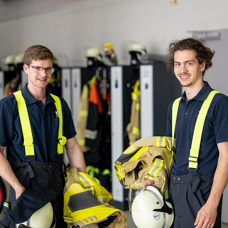 Zwei junge Personen stehen in einem Umkleideraum der Feuerwehr.