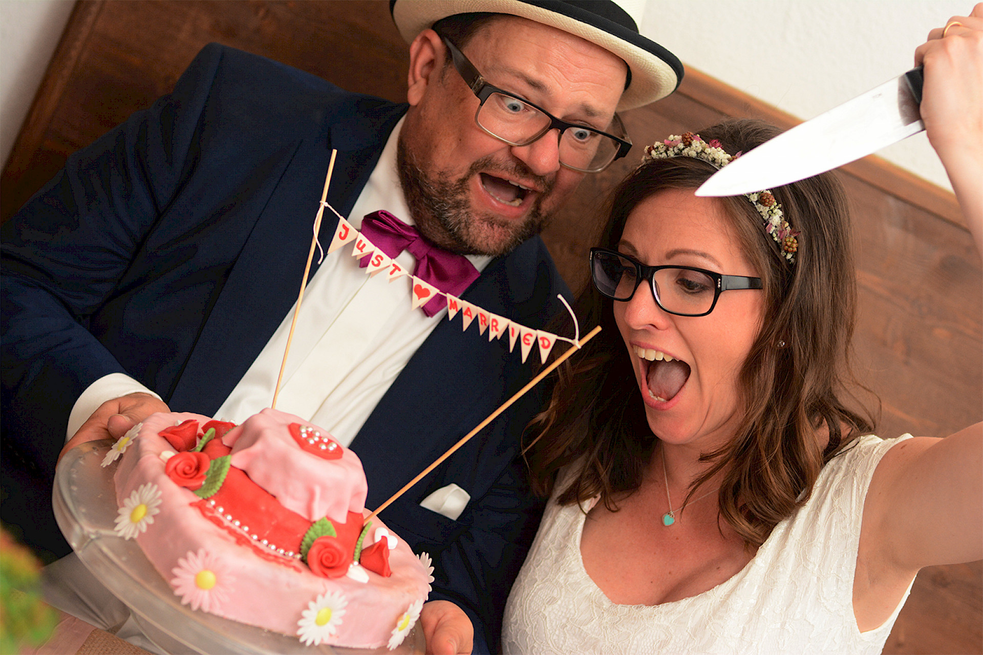 ein frisch verheiratetes Paar posiert humorvoll mit einer Hochzeitstorte. Der Bräutigam in Anzug und Hut hält die Torte mit der Aufschrift „Just Married“, während die Braut mit einem freudigen Gesichtsausdruck ein großes Messer über die Torte hebt. Beide wirken fröhlich und überrascht.