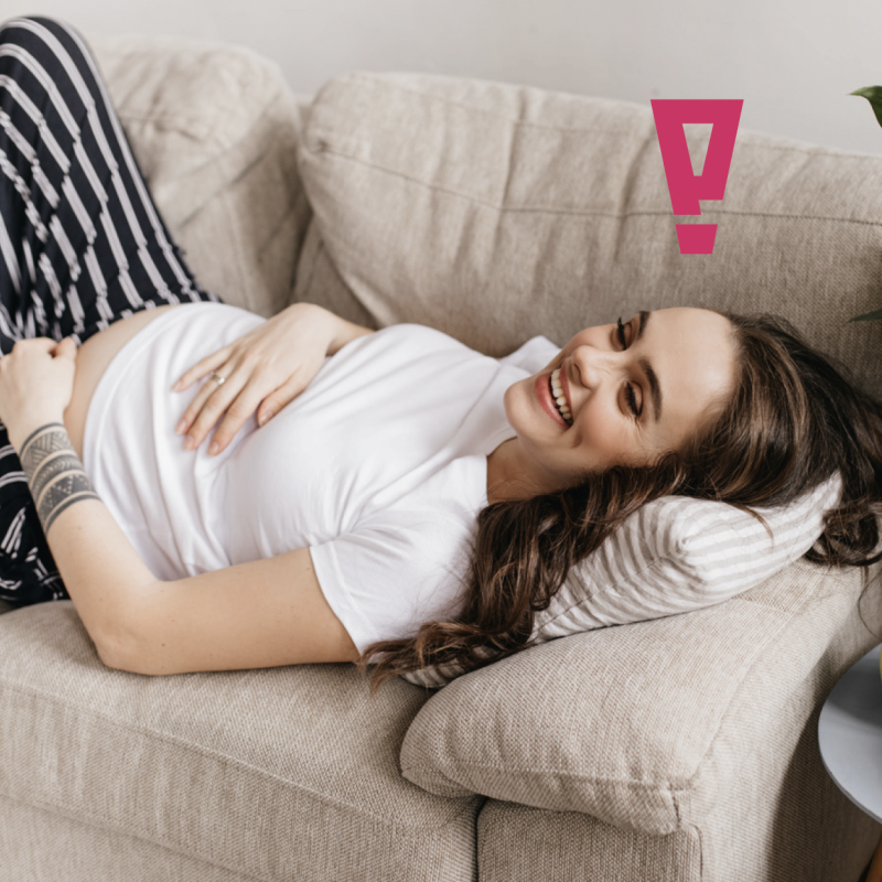 Schwangere Frau liegt lachend auf einer Couch und hat ihre Hände auf den Bauch gelegt.
