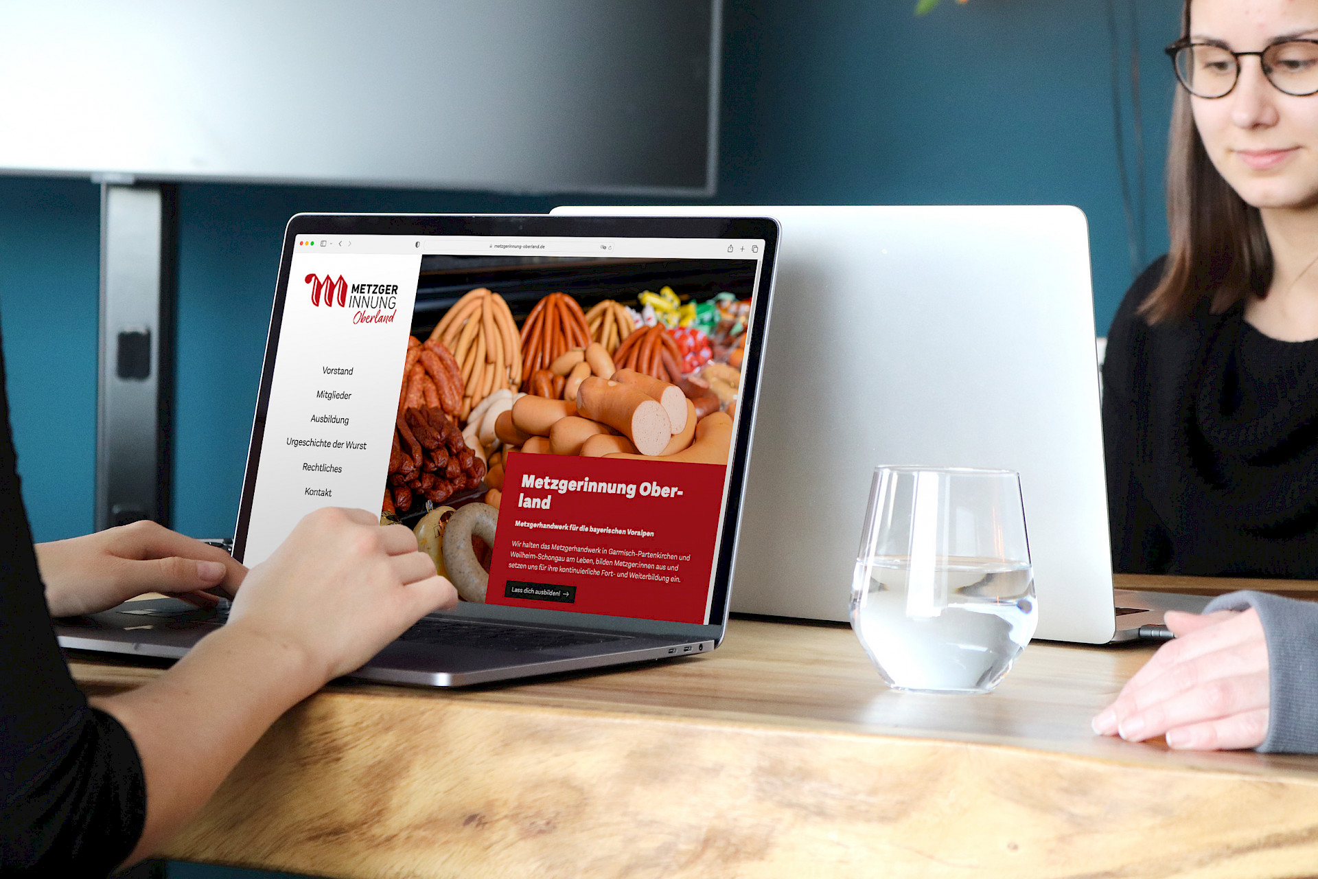 Zwei Personen sitzen an einem Tisch, der die Website der Metzgerinnung Oberland mit verschiedenen Wurstsorten anzeigt.