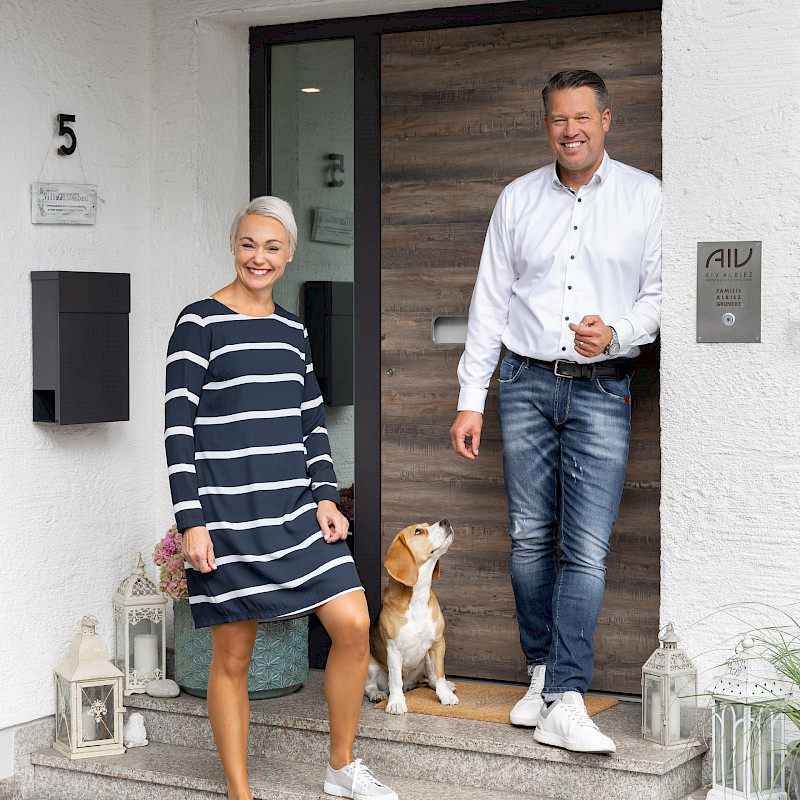 Frau und Mann stehen gemeinsam mit einem Hund freundlich lächelnd vor einer modernen Haustür